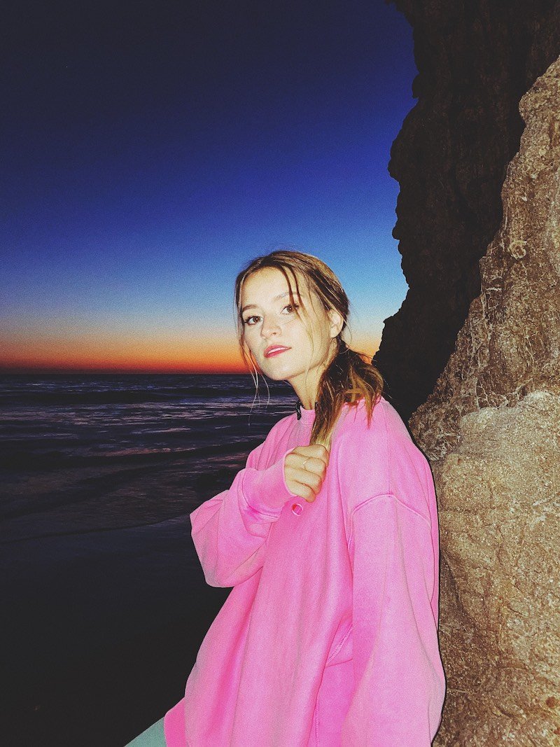 Rosie Darling press photo wearing a pink sweatshirt with a body of water in the background 