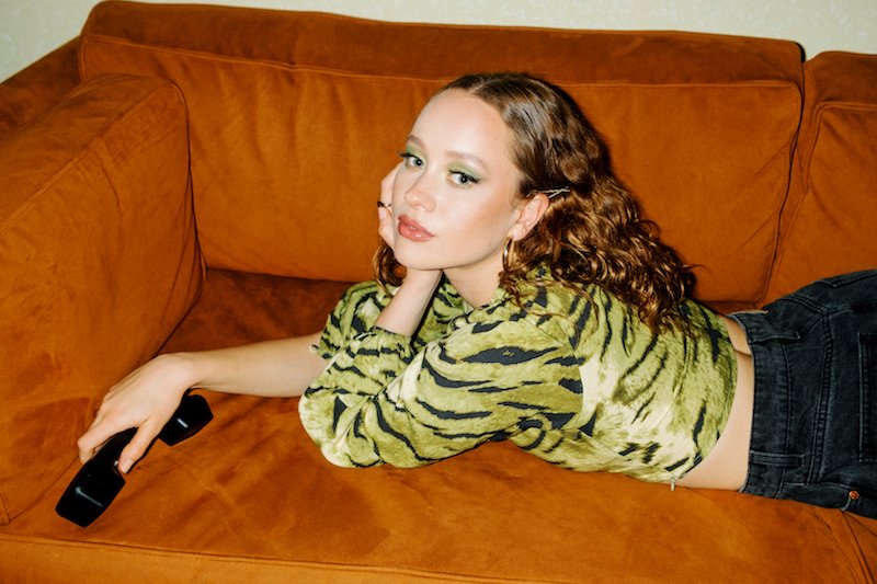 Piper 57 press photo wearing a black and yellow outfit while laying on an orange sofa