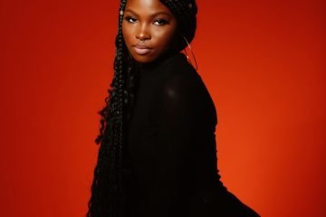 Ria Sean press photo wearing a black outfit with a red background