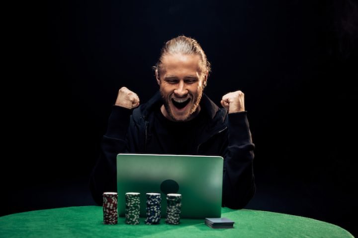 happy man gesturing while using laptop near poker chips isolated on black.