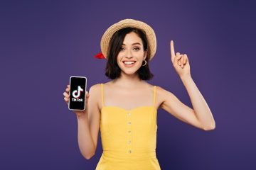 Excited girl in straw hat showing idea gesture and holding smartphone with tiktok app isolated on purple